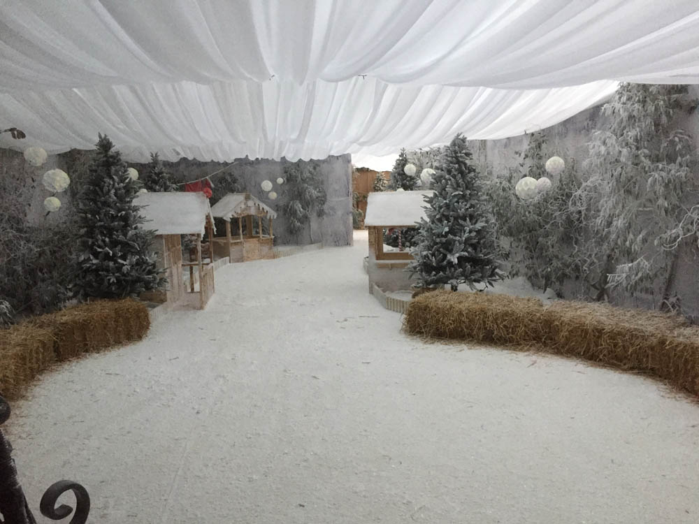 Interior of a Christmas marquee decorated as a Santa’s grotto with snowy flooring, hay bales, fir trees and festive lighting.