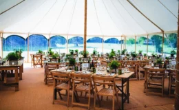 Open-sided pole marquee set for dining with wooden tables and chairs, providing shade for an outdoor event.