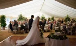 Bride and groom entering a lined wedding marquee with seated guests, floral arrangements and a wooden dance floor.