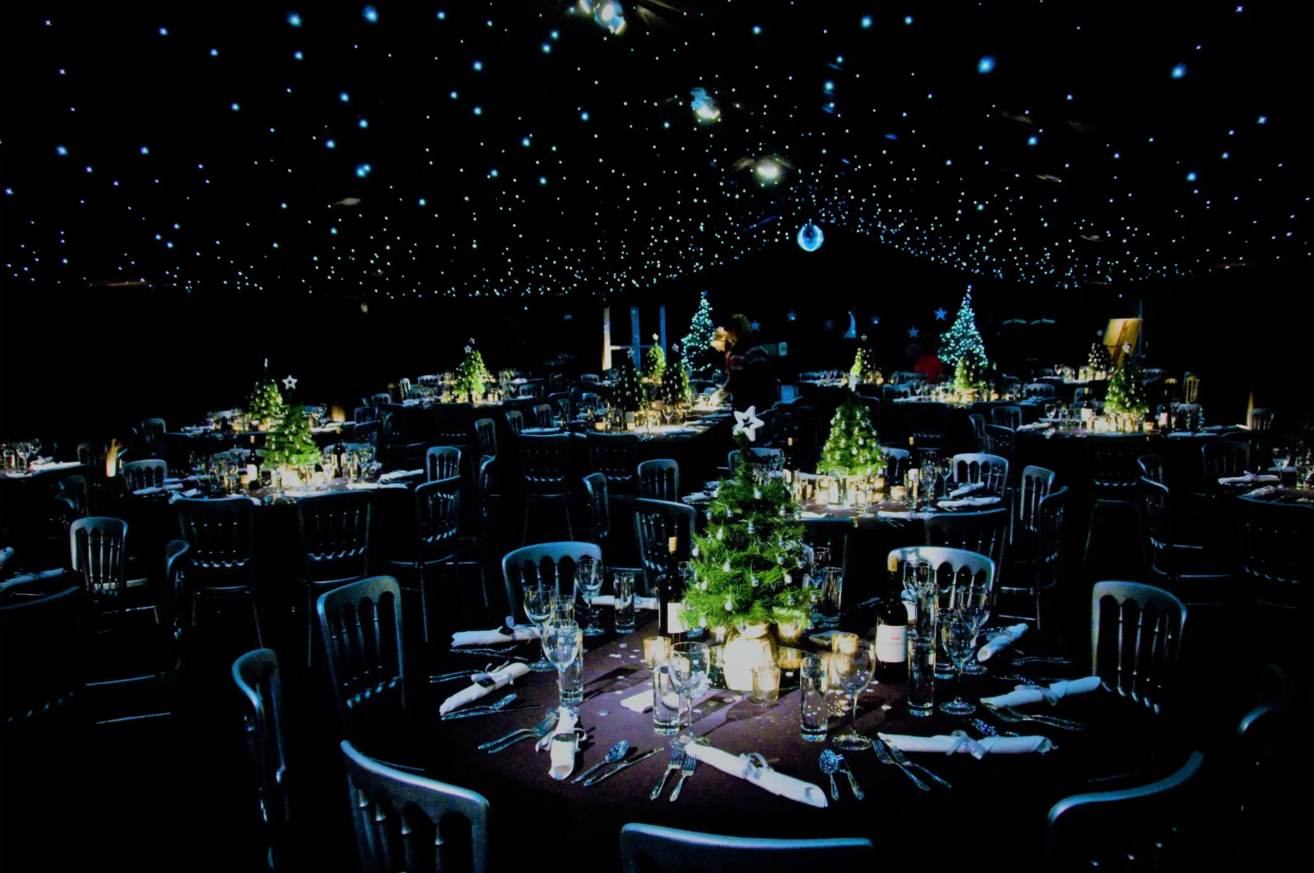 Blackout Christmas marquee interior with starlit LED ceiling and round tables decorated with mini Christmas trees.