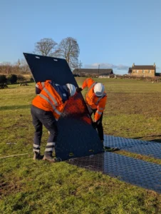 Two workers in high-visibility jackets and helmets installing interlocking HDPE trackway panels on a grassy field to create a temporary roadway